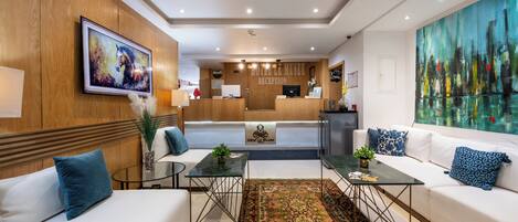 Un hall d’hôtel moderne avec un comptoir d’accueil, des canapés blancs, une grande peinture abstraite et un tapis à motifs traditionnels.