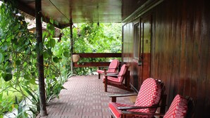 Une véranda couverte, avec des chaises en bois et un banc, entourée de verdure.