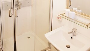 Une douche fermée par des parois en verre, dotée d’un pommeau de douche à main, un lavabo blanc avec un seul robinet et un miroir au-dessus du lavabo.
