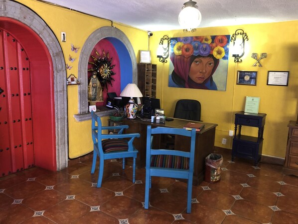 A room with a red door, a blue chair, a desk with a computer, and a painting on the wall. - Posada Margaritas (Tlaquepaque)