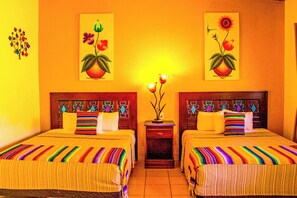A hotel room with two beds, colorful striped bedding, a nightstand with a vase of flowers, and two wall-mounted paintings of flowers.