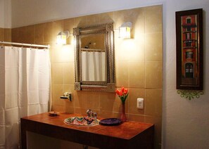 A bathroom with a tiled wall, a mirror with ornate frame, a sink with a colorful mat, a vase with flowers, and a framed picture on the wall.
