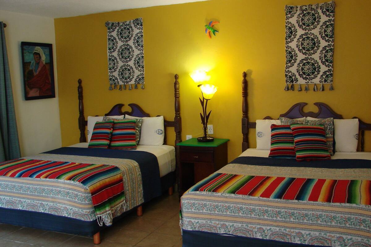 A room with two beds, each with colorful striped bedding, a nightstand with a lamp, and a wall hanging above the beds.
