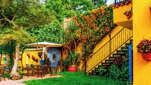 A courtyard with a yellow building, outdoor seating, and a staircase. - Posada Margaritas (Tlaquepaque)