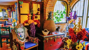 A room with a wooden chair, a red suitcase, and a vase of flowers. - Posada Margaritas (Tlaquepaque)