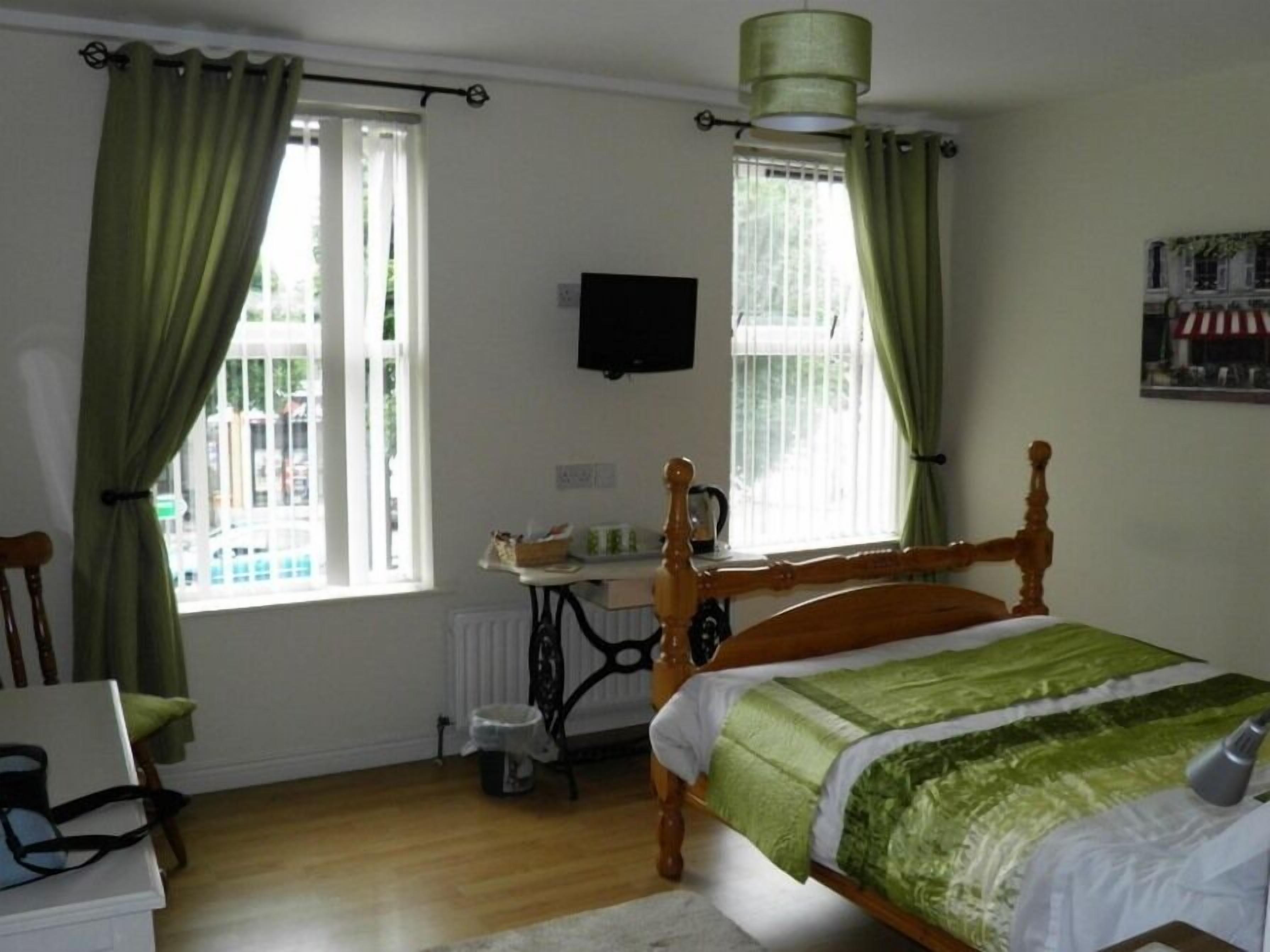 Quarto com cama de madeira, cortinas verdes, radiador, escrivaninha com cadeira e televisão na parede.