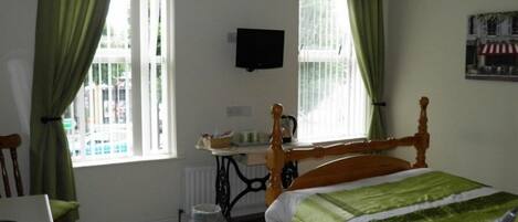 Quarto com cama de madeira, cortinas verdes, radiador, escrivaninha com cadeira e televisão na parede.