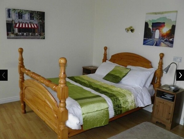 A wooden four-poster bed with a green and white bedspread, a nightstand with a lamp, and two wall-mounted paintings.