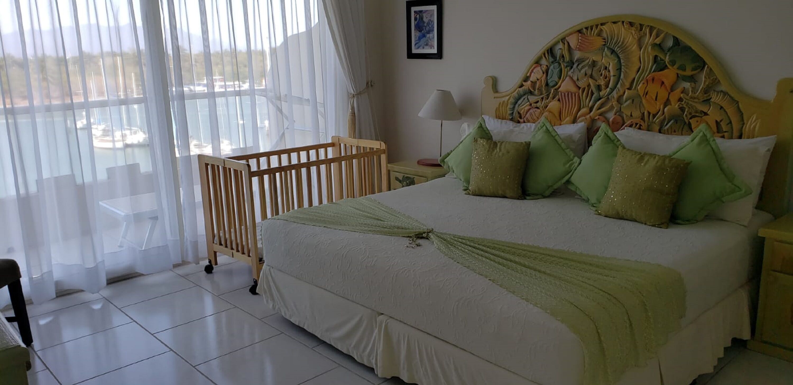A hotel room with a bed, a crib, a window with curtains, a lamp, and a decorative headboard.