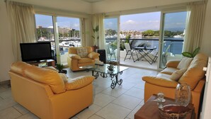 Un salón amplio con un sofá grande, una mesa de centro y un televisor. La habitación tiene ventanas grandes con vistas a un puerto deportivo y edificios.