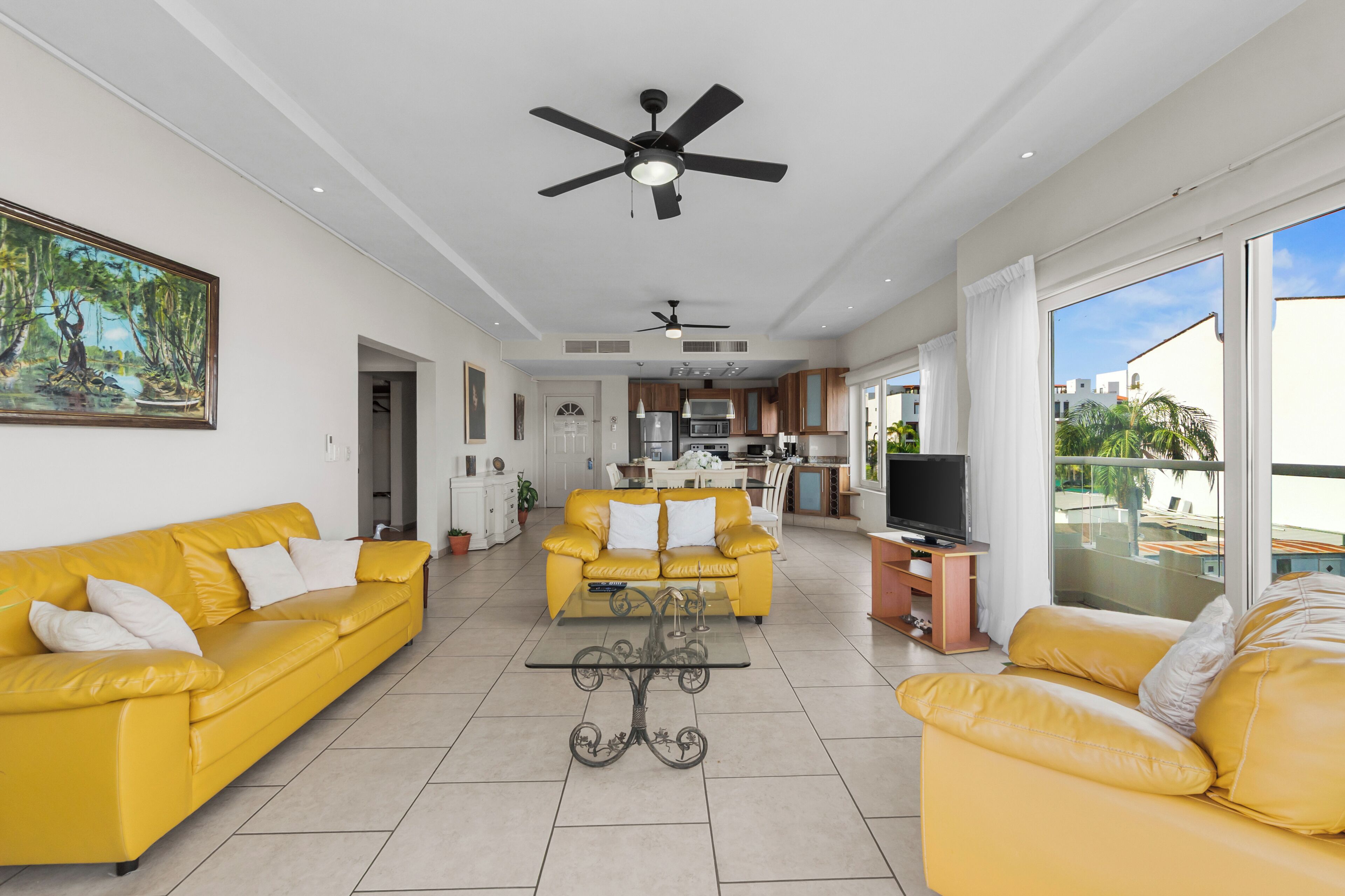 A spacious living room with yellow sofas, a glass coffee table, and a TV on a wooden stand.