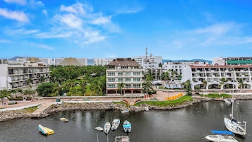 Un hotel frente al mar con varios balcones, un puerto deportivo con varios barcos y edificios urbanos al fondo.