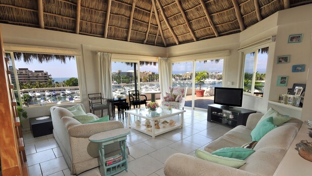 Un salón amplio con un sofá grande, una mesa de centro y un televisor. La habitación tiene ventanas grandes con vistas a un puerto deportivo y edificios.