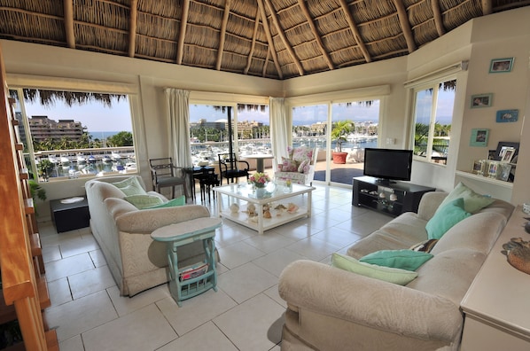 Un salón amplio con un sofá grande, una mesa de centro y un televisor. La habitación tiene ventanas grandes con vistas a un puerto deportivo y edificios.