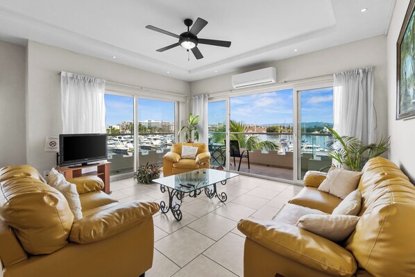 A spacious living room with a flat-screen TV, two sofas, a glass coffee table, and a ceiling fan. - Marina Banderas Suites (Nuevo Nayarit)