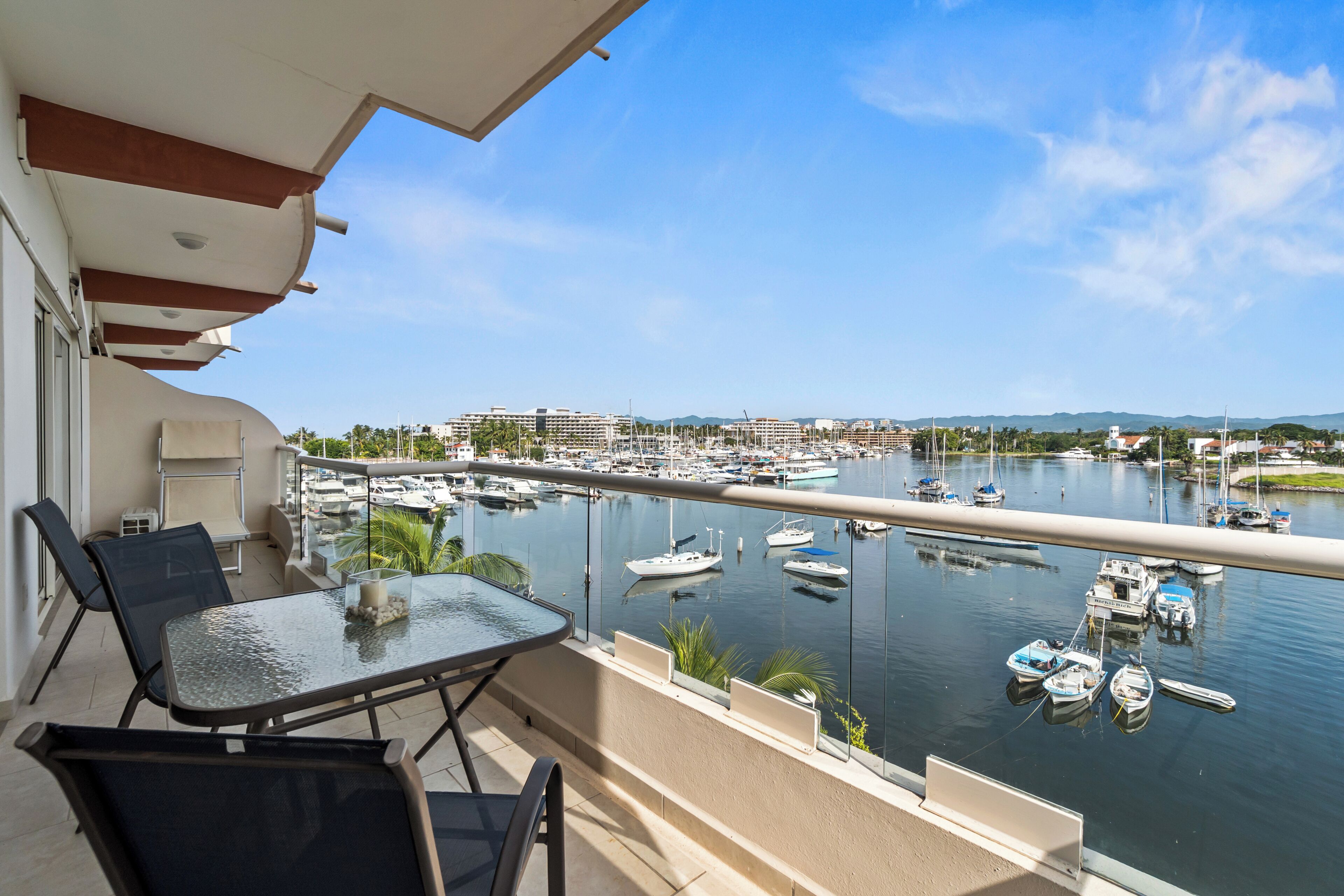 A balcony with a glass table and chairs overlooking a marina with boats and buildings in the background.