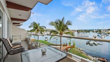 A balcony with a glass table and chairs, overlooking a marina with boats and palm trees.