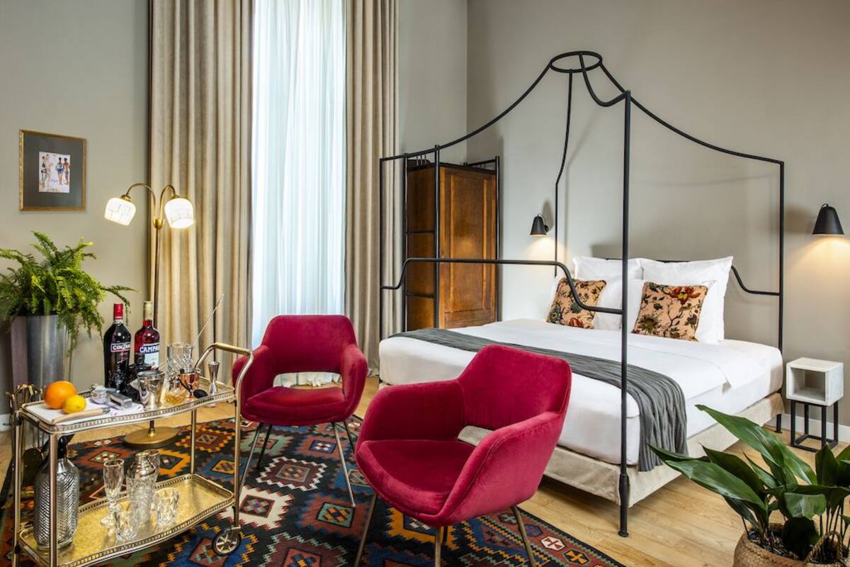 A hotel room with a four-poster bed, a red chair, a small table with drinks, and a patterned rug.