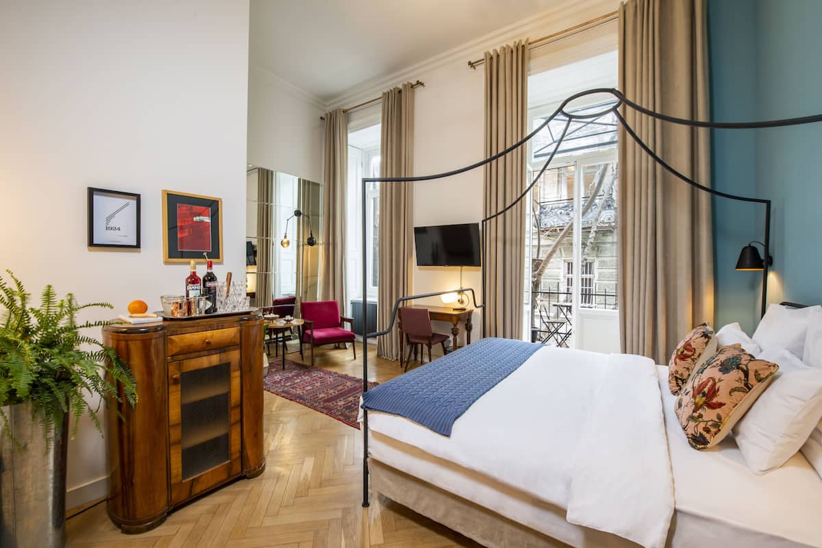 A hotel room with a large bed, a desk, a chair, a TV, and a view of a building outside.
