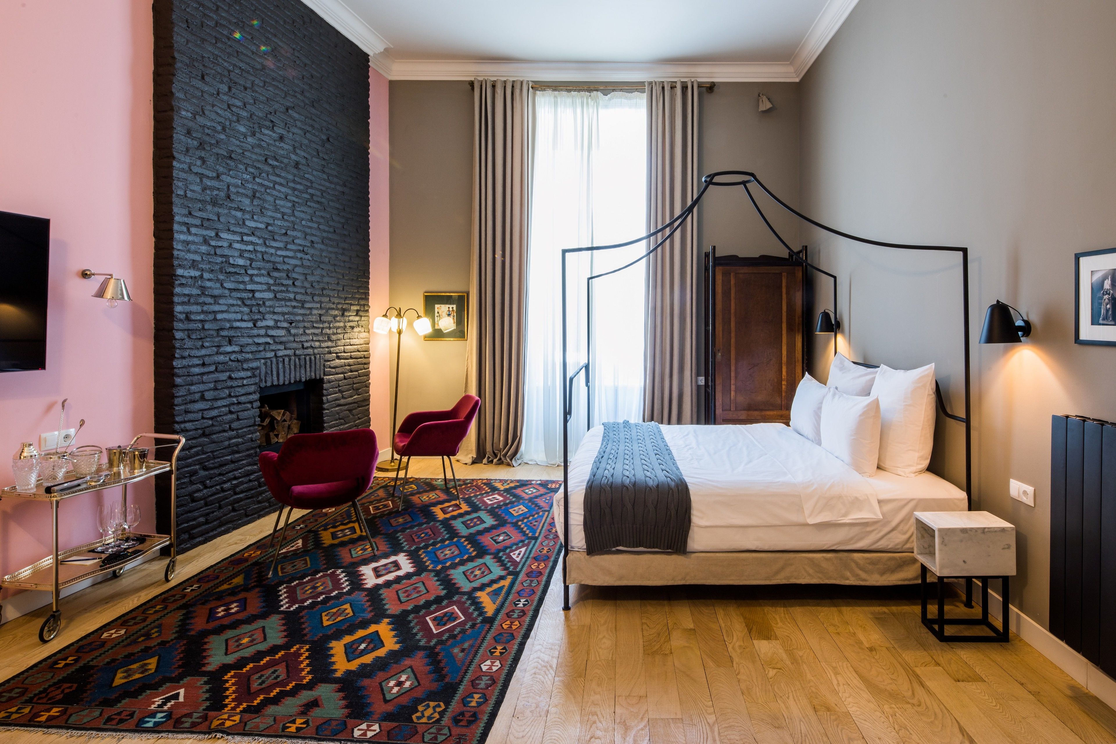 A modern bedroom with a brick wall, a large bed, a red chair, a patterned rug, and a small table with glasses.