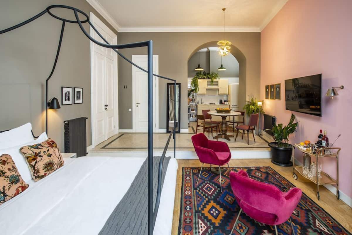 A bedroom with a bed, a red chair, a patterned rug, and a kitchen in the background.