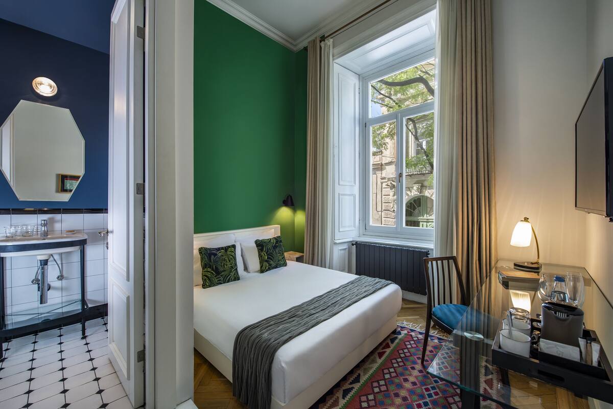 A hotel room with a bed, a glass table, a chair, a window with curtains, a radiator, and a bathroom with a mirror and sink.