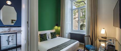 A hotel room with a bed, a glass table, a chair, a window with curtains, a radiator, and a bathroom with a mirror and sink.