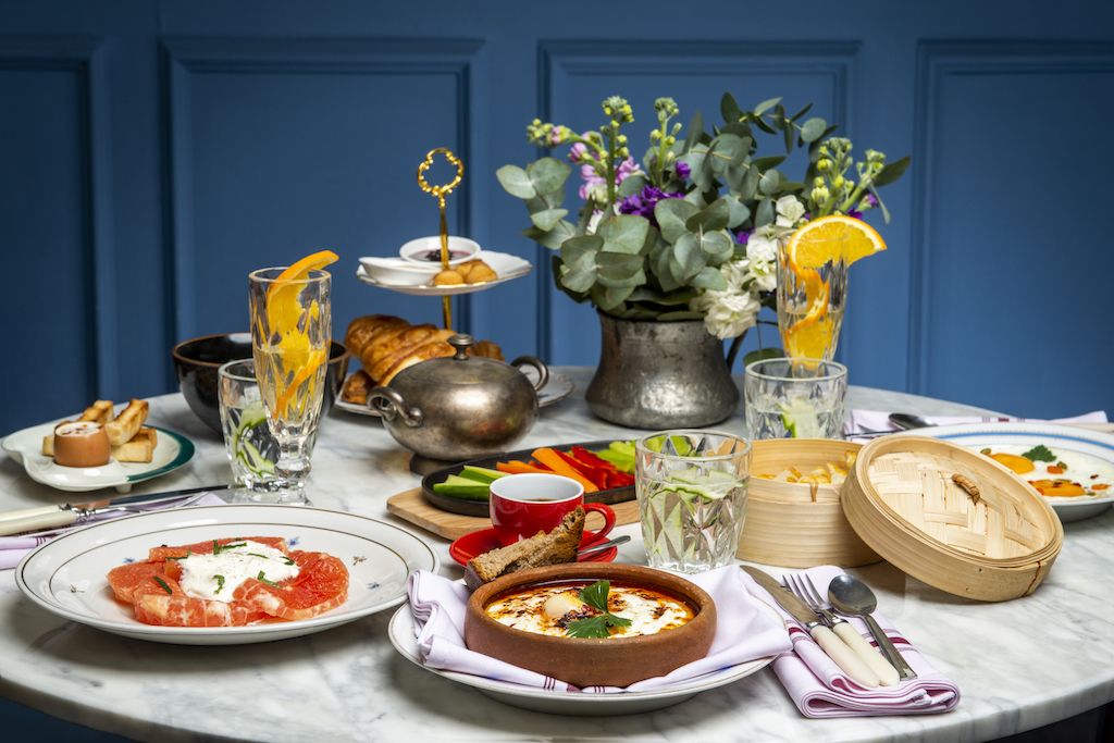 Ein Tisch ist reichhaltig gedeckt, mit verschiedenen Gerichten, darunter Bruschetta, eine Suppe und Obstsalat. Mehrere Gläser Wasser und eine Vase mit Blumen gehören ebenfalls dazu.