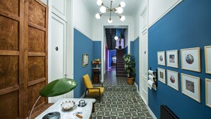 Un pasillo con paredes azules, piso estampado, una silla verde, una mesa con lámpara y cuadros en la pared.
