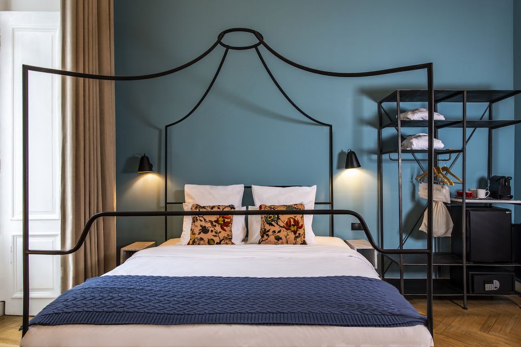 Un lit avec un cadre en métal, des draps blancs et une couverture bleue. On aperçoit une étagère avec des vêtements et une machine à café à l’arrière-plan.