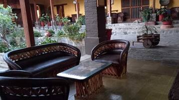 A covered outdoor seating area with wicker furniture, potted plants, and a stone floor.