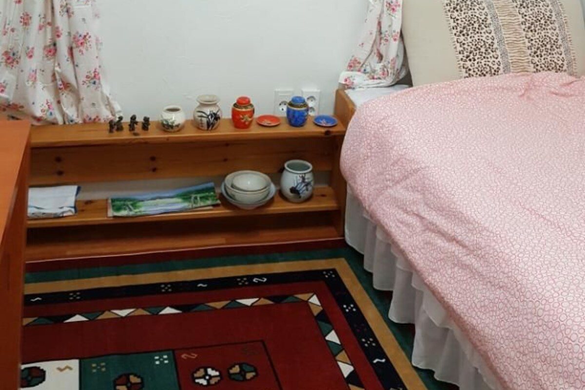 A bedroom with a window, a bed, a bedside table with decorative items, and a patterned rug.