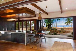 A modern kitchen with a large island, bar stools, and a view of the beach. - Spice At Oak Absolute Oceanfront House (Oak Beach)