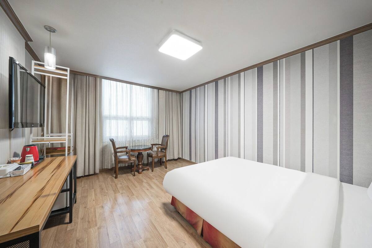 A hotel room with a bed, a desk with a chair, a television, and a window with curtains.