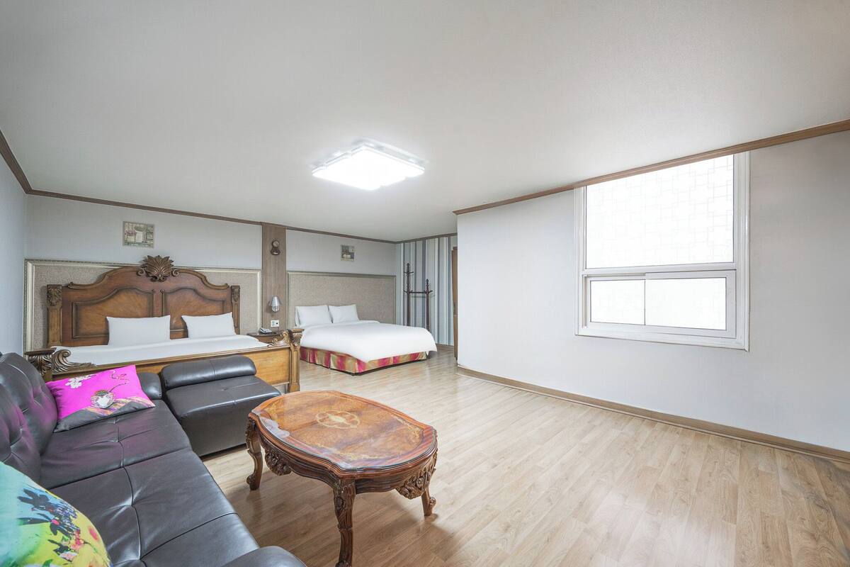 A hotel room with a large bed, a sofa, a wooden coffee table, and a window providing natural light.