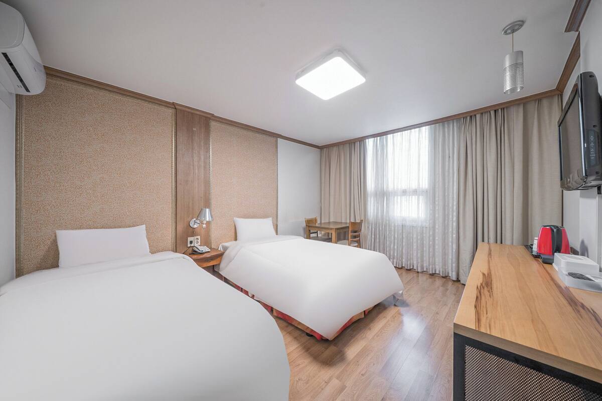 A hotel room with two beds, a wooden desk, a television, and a window with curtains.