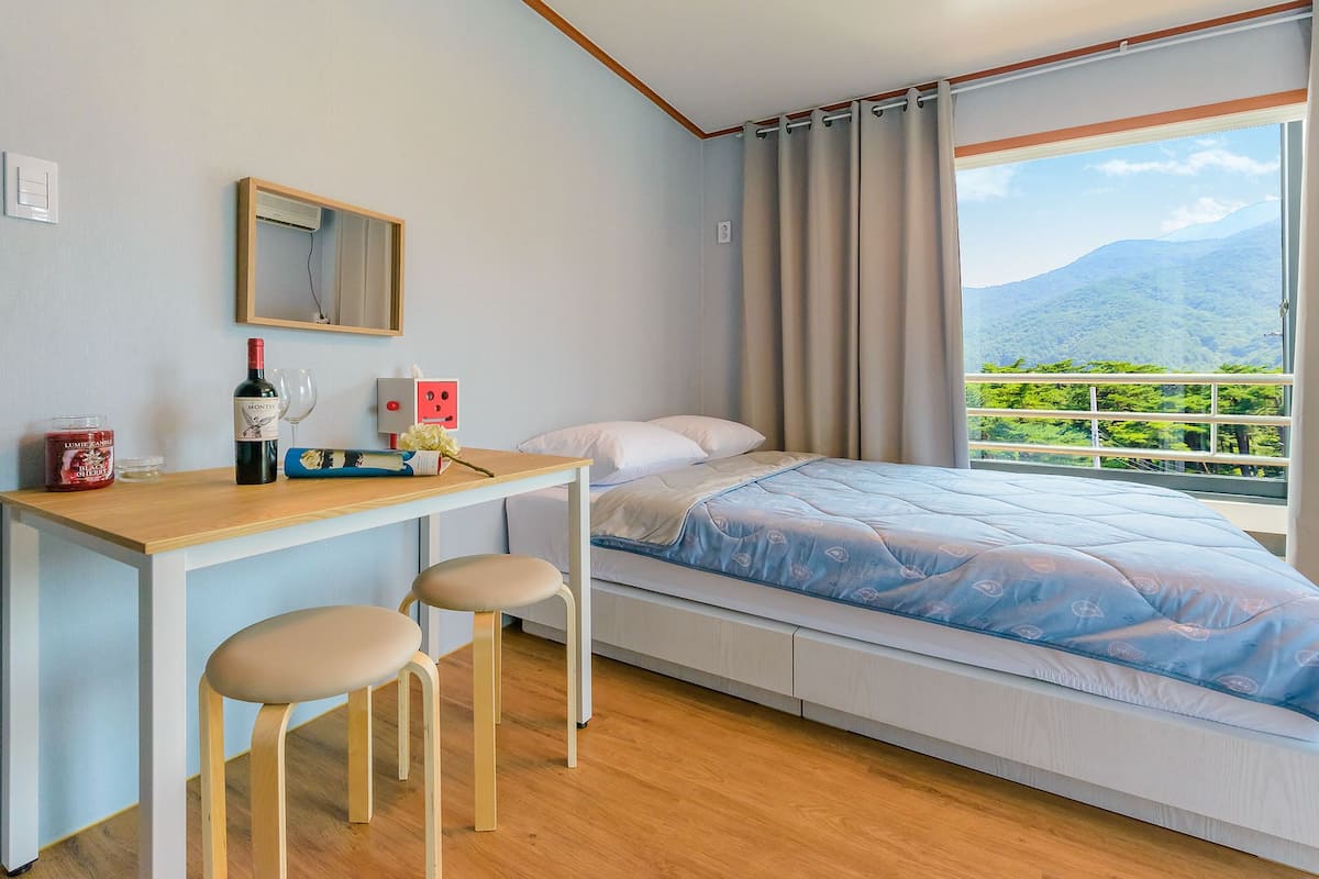 A bedroom with a bed, a desk with two stools, a mirror, and a view of a mountainous landscape through a window.