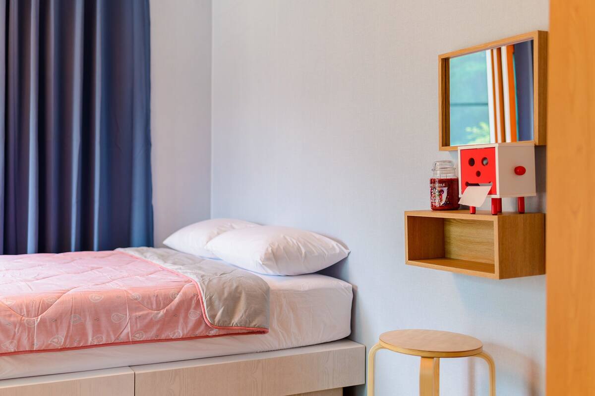 A bedroom with a bed, a bedside table, a small stool, a shelf with a candle and a toy, and a window with curtains.