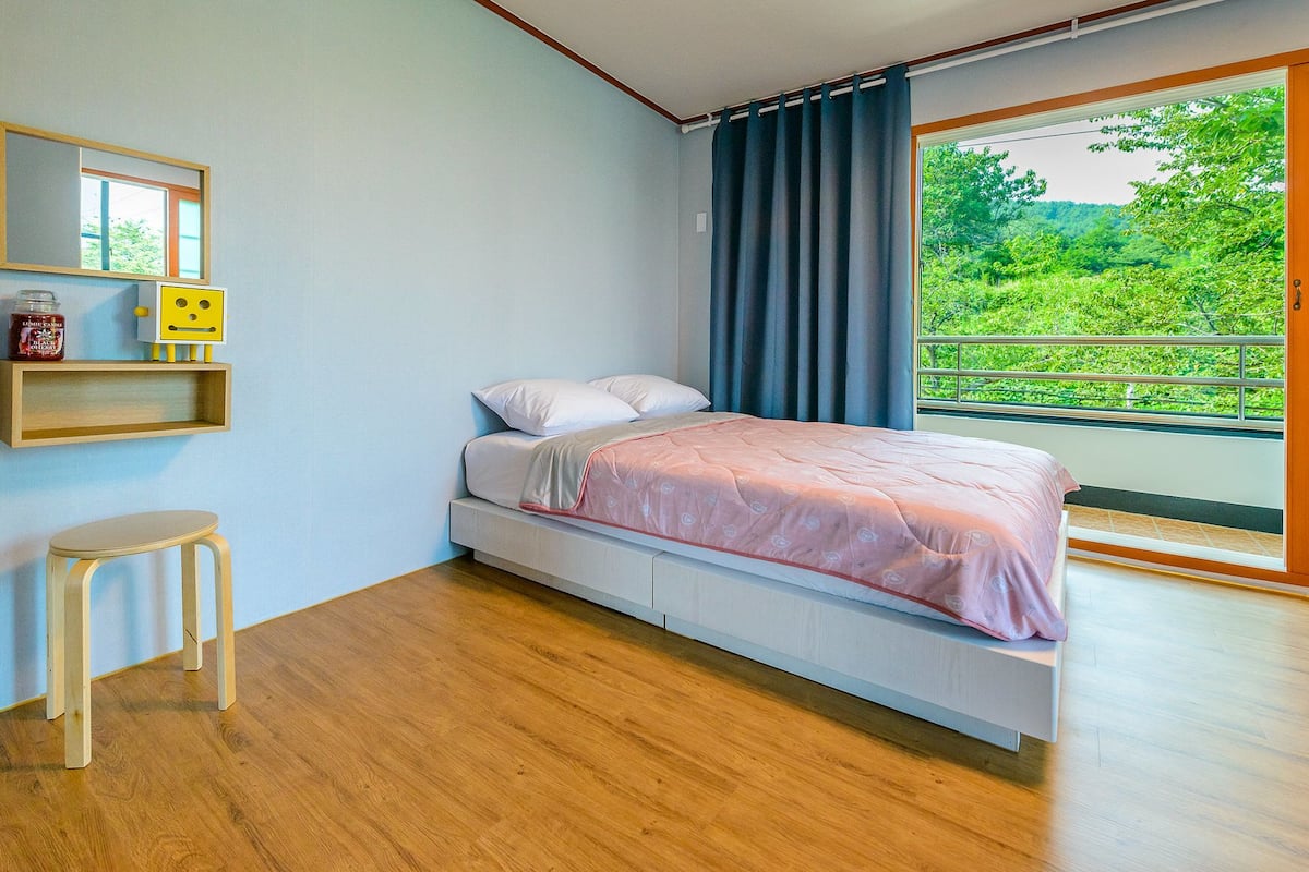 A bedroom with a bed, a small table, a shelf with items, a mirror, and a large window with a view of greenery.
