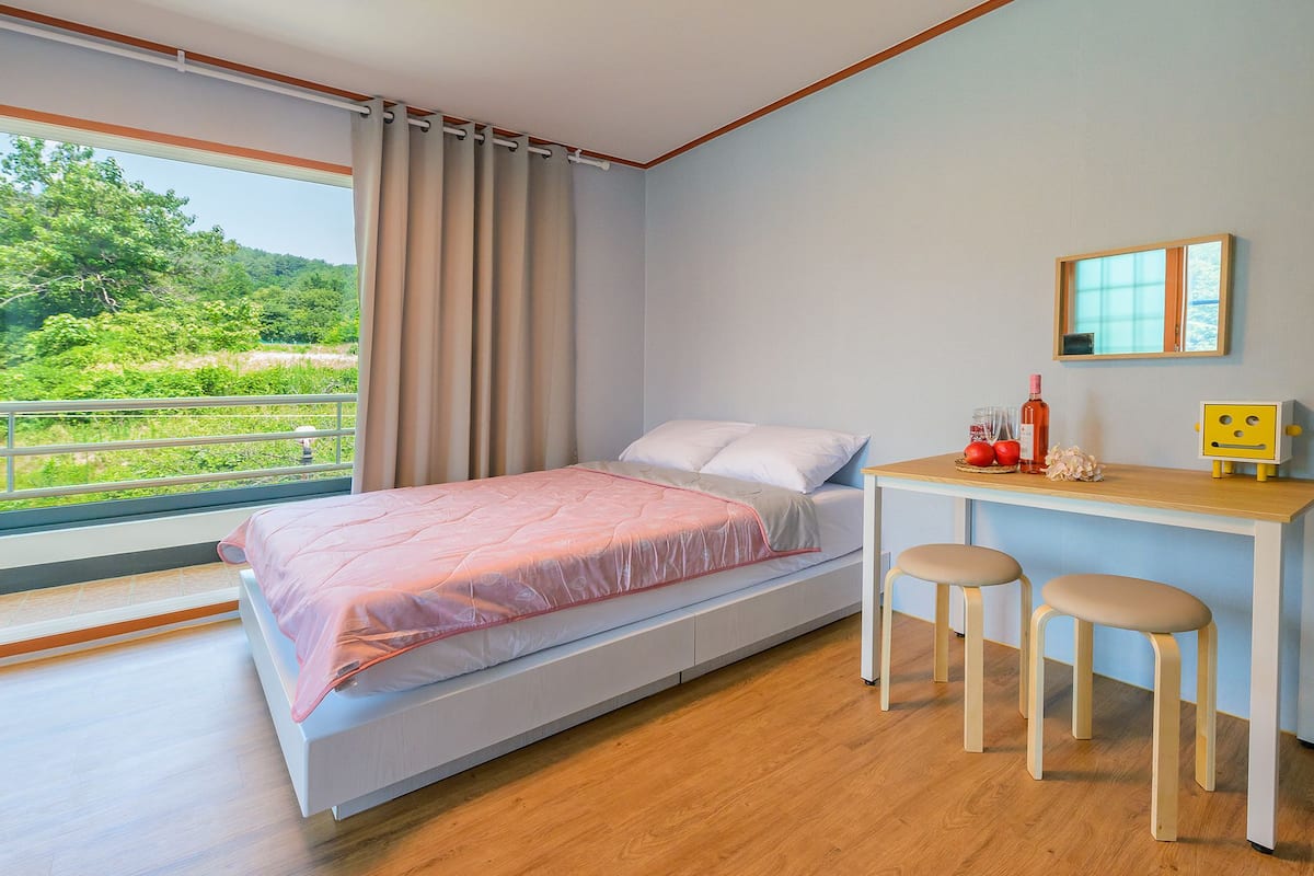 A bedroom with a bed, a desk, two stools, a mirror, and a view of greenery outside.