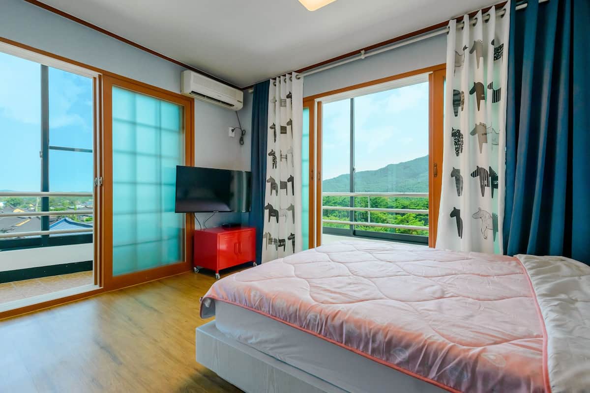 A bedroom with a large bed, a TV on a stand, and a balcony with a view of greenery and mountains.