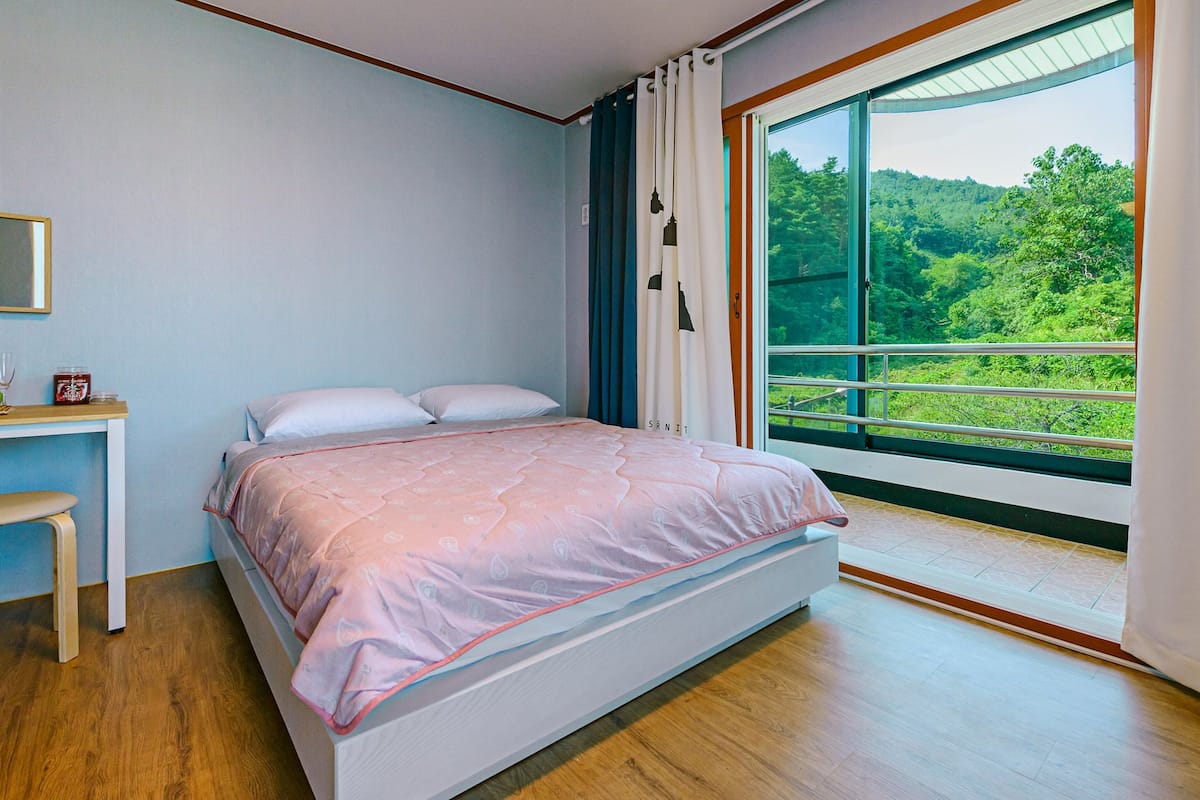 A bedroom with a bed, a desk, and a large window with a view of greenery.