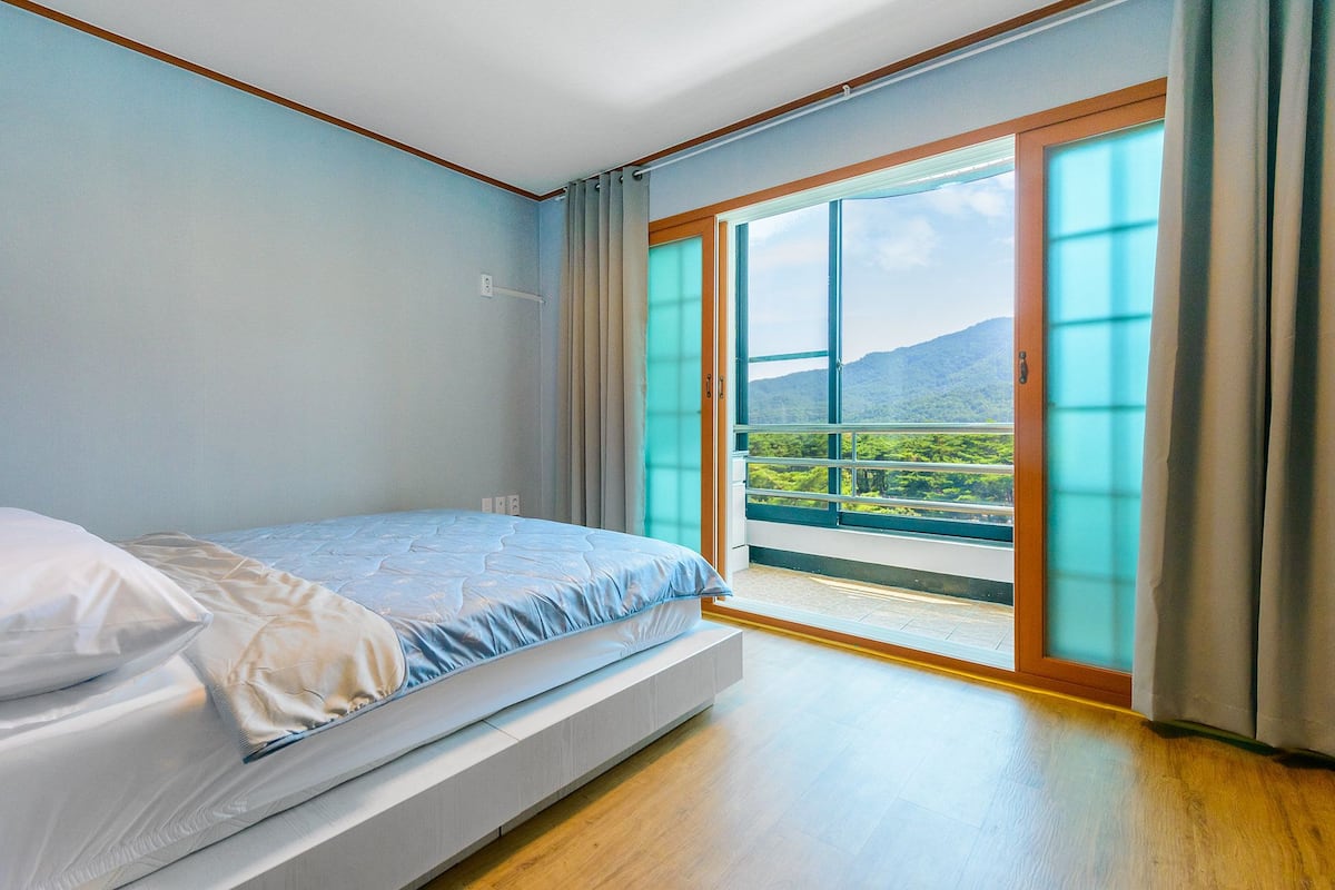 A bedroom with a bed, a window with curtains, and a view of greenery and mountains.