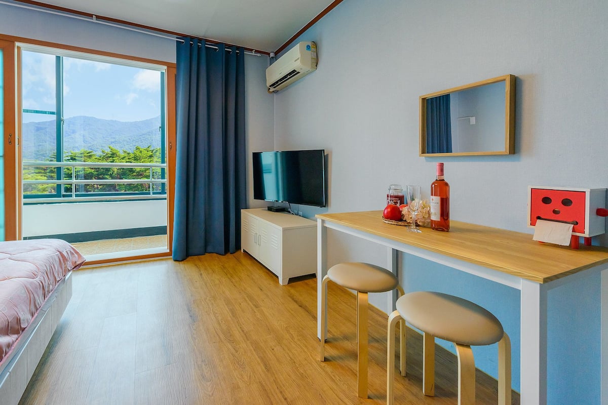 A hotel room with a bed, a desk with two chairs, a TV, a small table with a bottle and glass, and a window with a view of greenery and mountains.