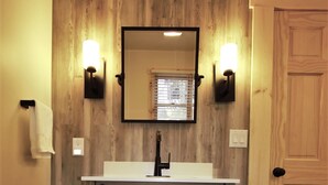 A bathroom with a wooden wall, a white sink, two wall-mounted lights, a mirror, and a door. - Beautifully Wooded & Serene Setting (Hot Tub) (Maggie Valley)