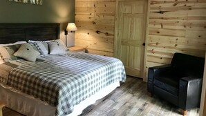 A bedroom with a bed, a nightstand, a lamp, a wooden door, and a black chair. - Beautifully Wooded & Serene Setting (Hot Tub) (Maggie Valley)