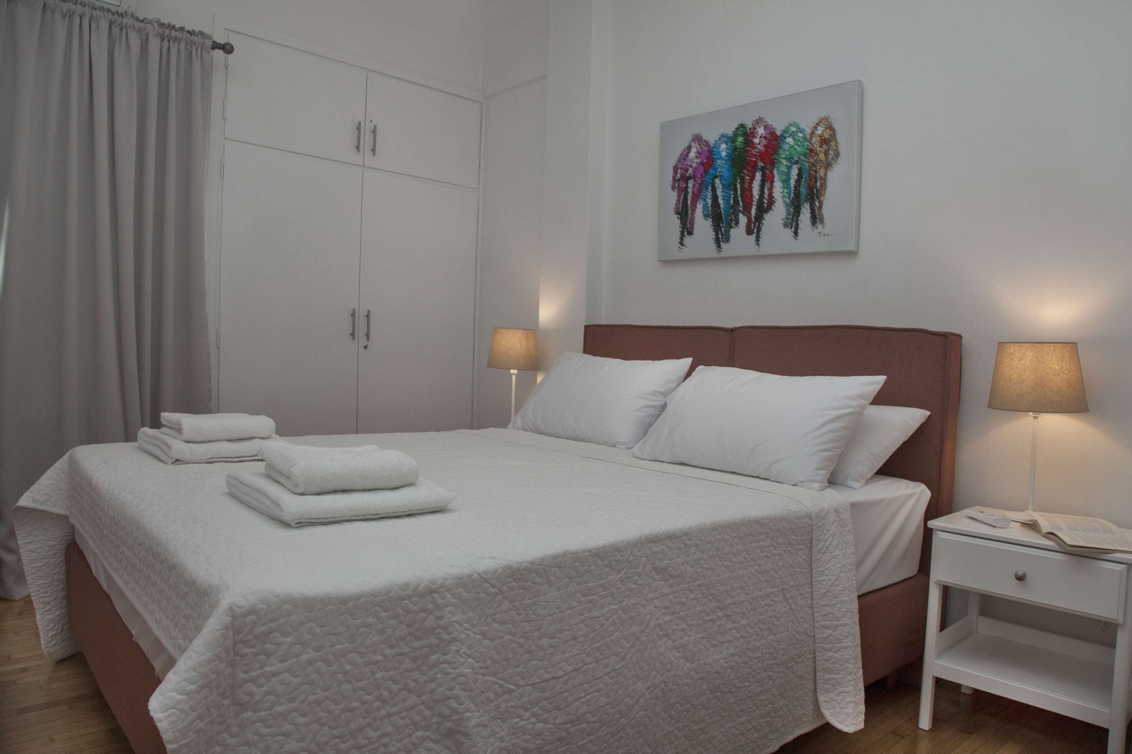 Een net opgemaakt bed met witte lakens en kussens, een nachtkastje met een lamp, en een aan de muur hangend kunstwerk.
