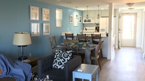 Una sala de estar con sofá, una mesa de centro con una botella y flores, un comedor con sillas y una mesa, y una cocina al fondo.
