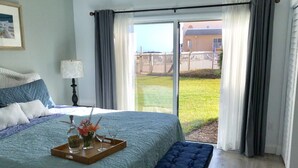 Un dormitorio con una cama grande, un banco, una bandeja con flores y vistas a un jardín césped a través de una puerta corrediza.