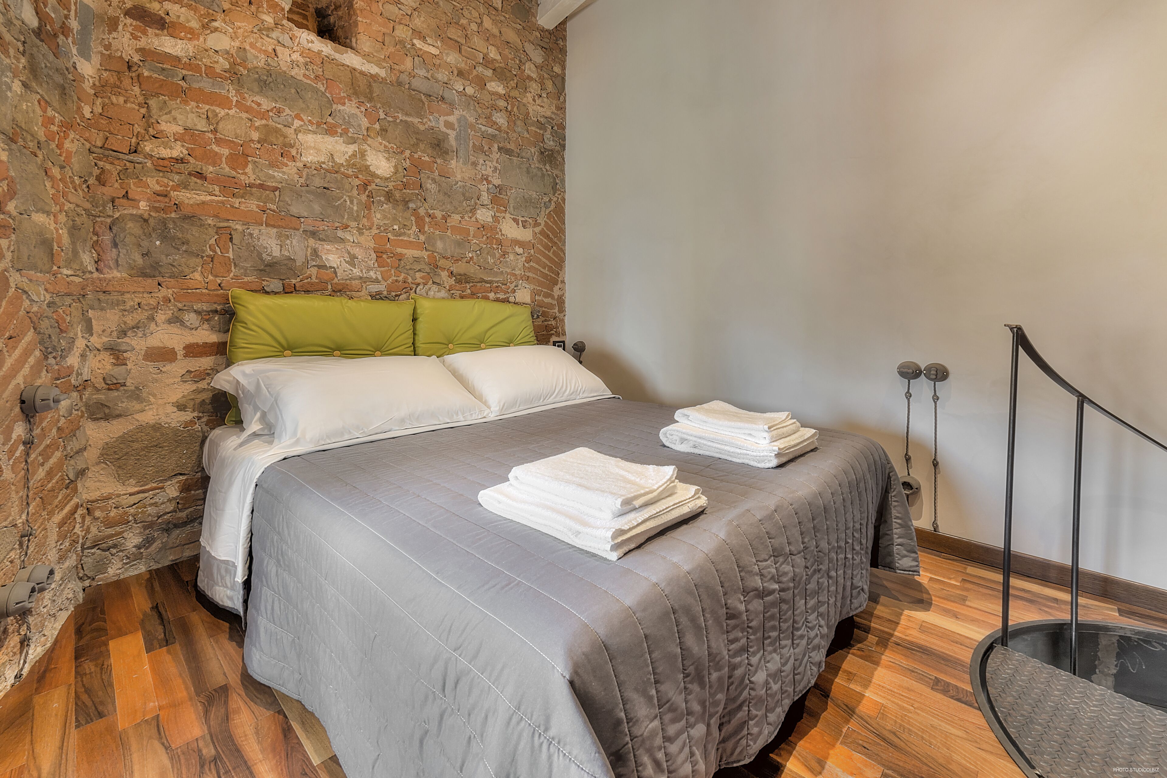 A room with a brick wall, a bed with grey bedding, and a wooden floor.